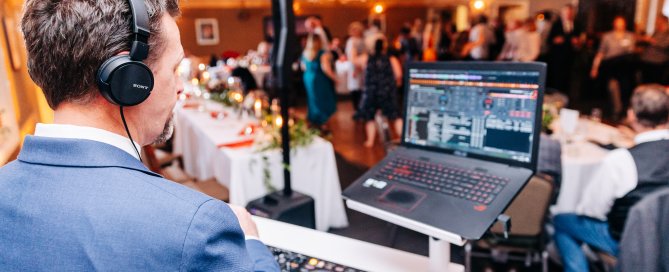 Wedding DJ in headphones mixing at the booth as guests dine behind him at an Alaska reception